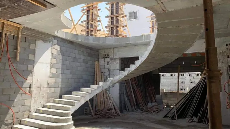 درج خرساني مسلح دائري مصبوب في الموقع يلتف صاعدًا نحو فتحة بيضاوية في البلاطة، بين جدران البلوك ودعامات الشدات